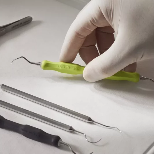 Main gantée en blanc, qui se saisit d'un outil dentaire jaune-vert, sur une surface avec d'autres outils dentaires. Fabriqué par Deppeler, entreprise certifiée B Corp suisse.